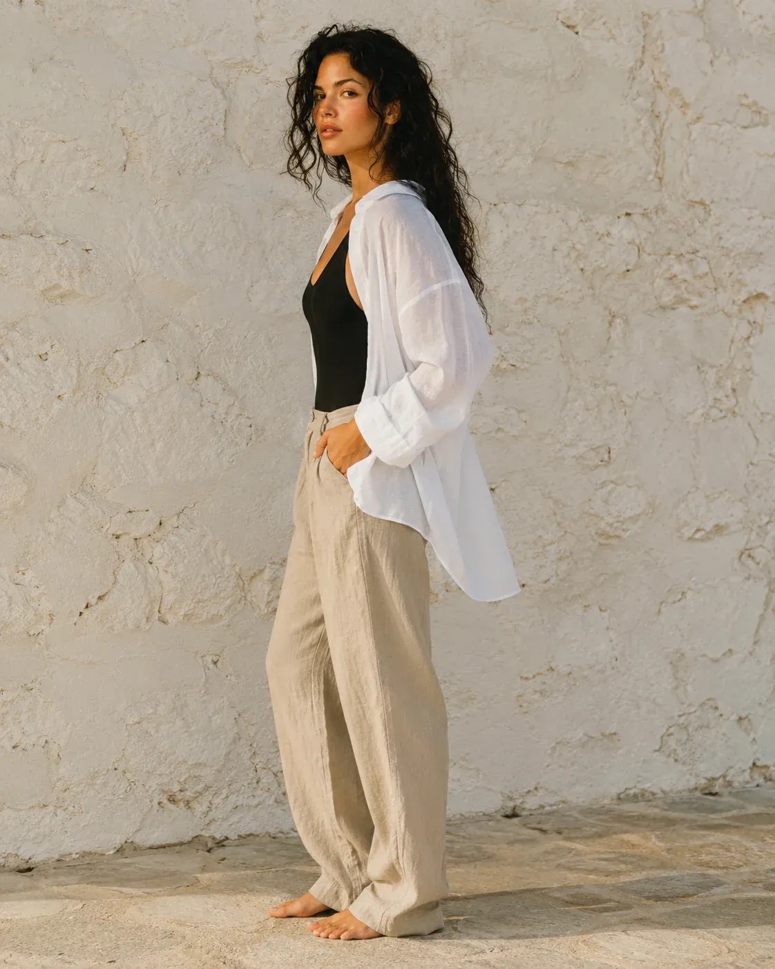 A woman in a white shirt and linen trousers standing sideways by a stone wall.