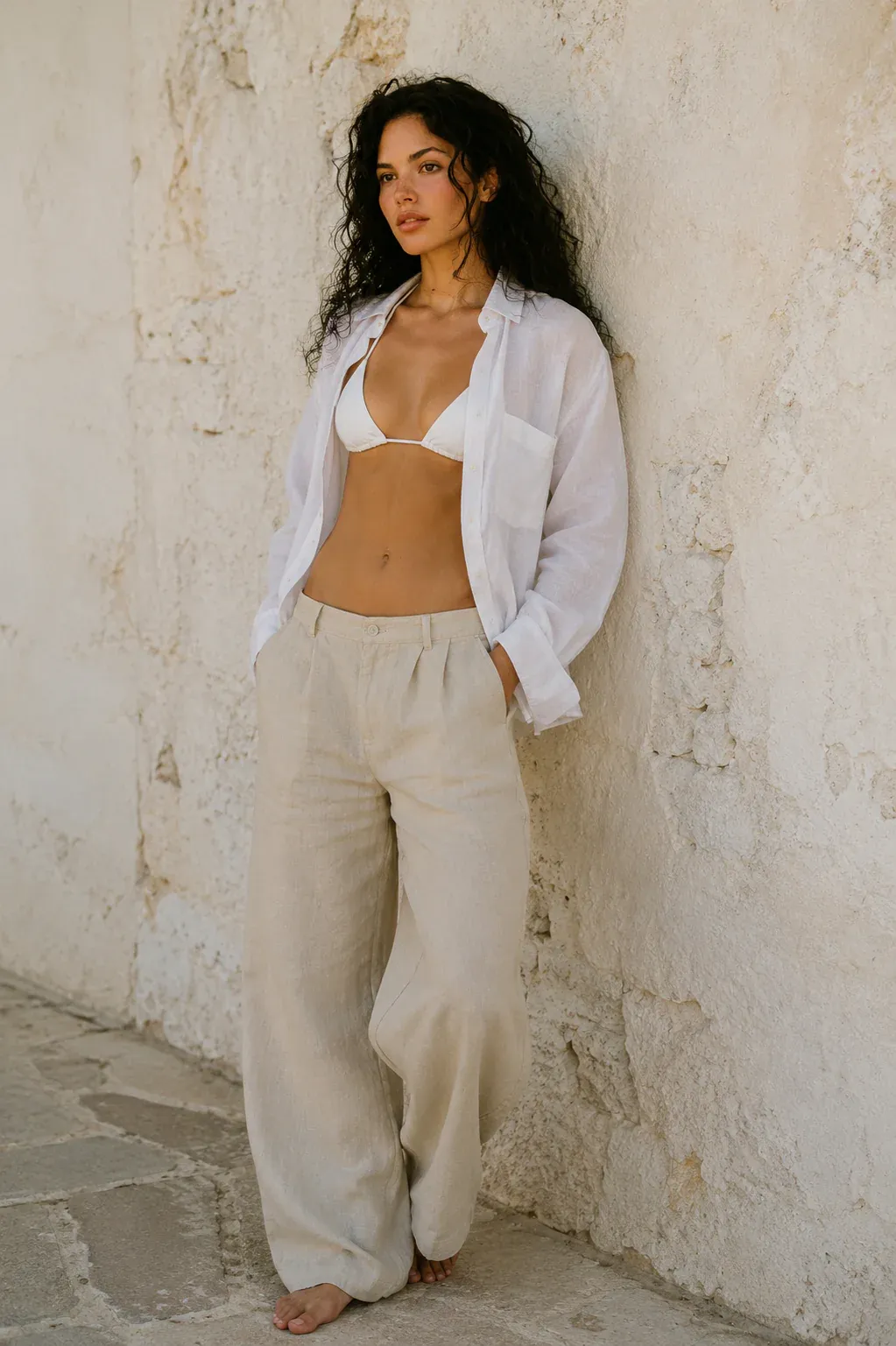 A woman in light linen clothing leaning against a stone wall.